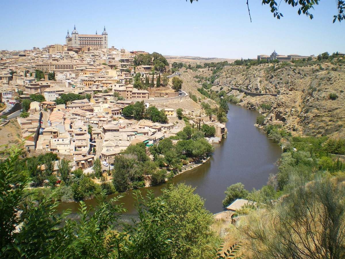 A hillside town with numerous buildings and greenery by a river, with a large historic building at the top, under a clear sky.