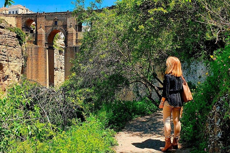Una persona de pie en un sendero rodeado de vegetación, contemplando un gran puente de arco de piedra en un día soleado.
