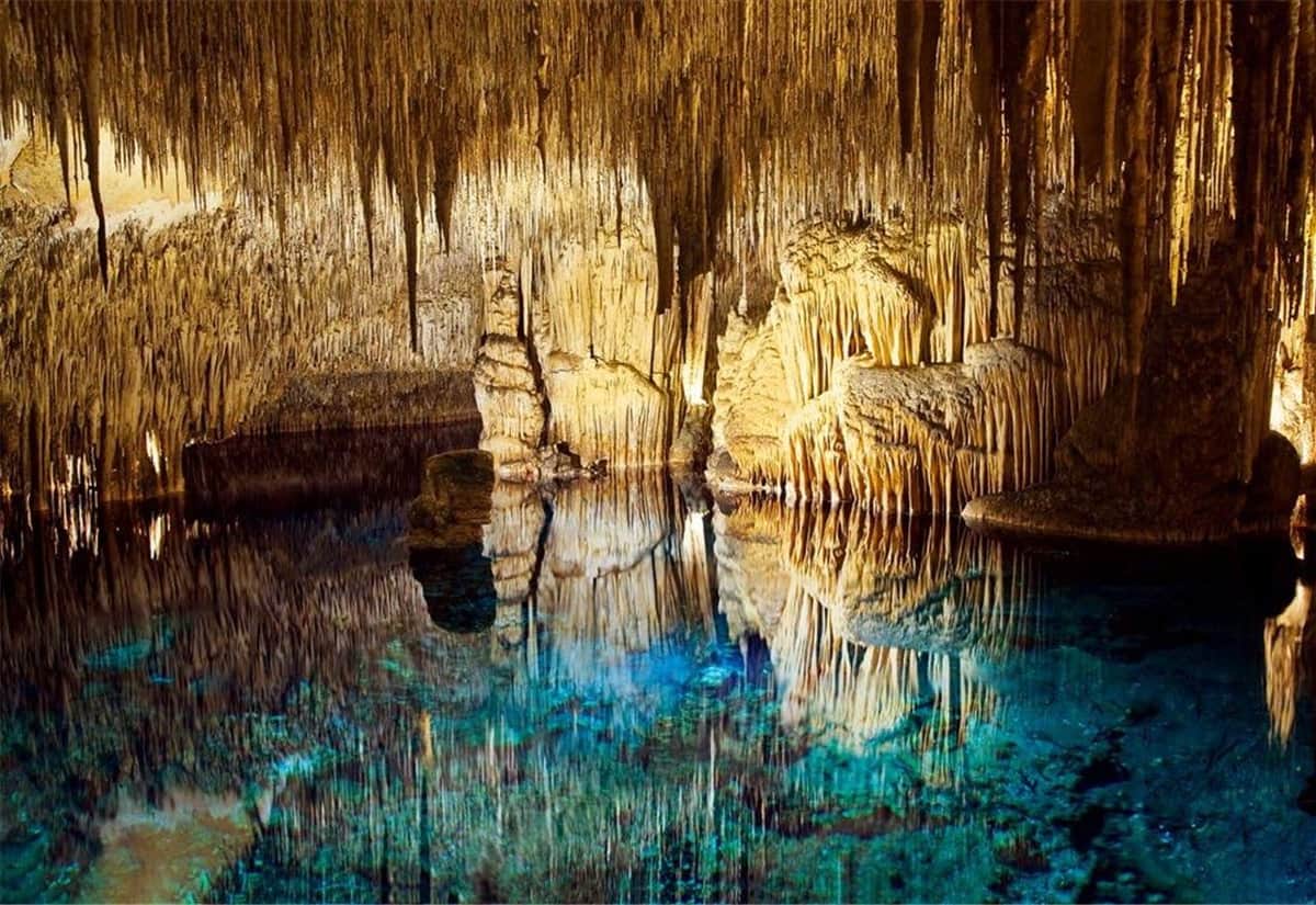 Une grotte avec des stalactites et des stalagmites illuminées qui se reflètent dans une piscine souterraine bleue et transparente.