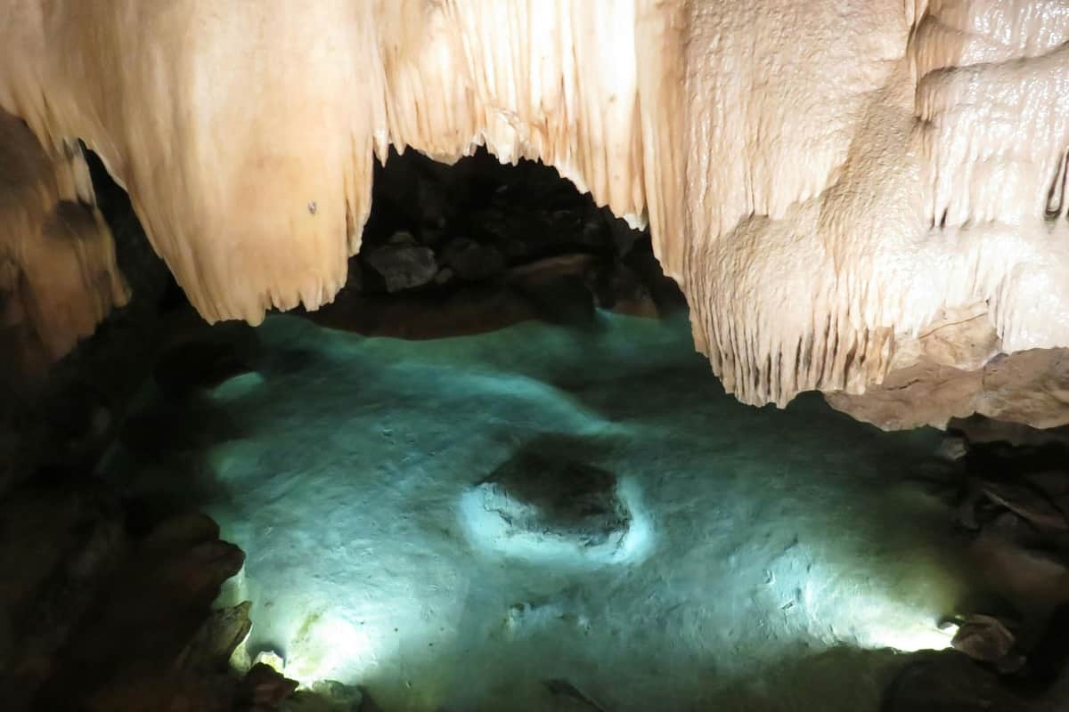 Des stalactites sont suspendues au-dessus d'une grotte faiblement éclairée, à l'eau turquoise claire et aux rochers immergés.