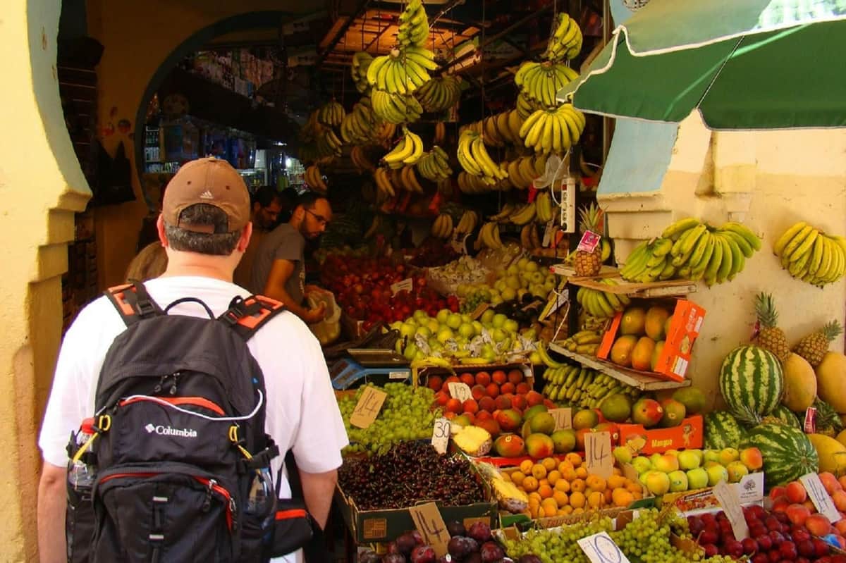 Un hombre con una mochila recorre un colorido mercado de frutas, con plátanos colgando y diversas frutas como manzanas, uvas y mangos.