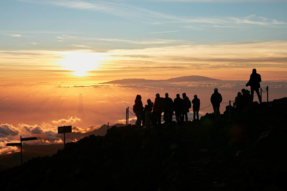 Mont Teide : Excursion au coucher du soleil, téléphérique + pique-nique Silhouette de personnes se tenant sur un chemin rocailleux au lever du soleil, surplombant une mer de nuages et des montagnes lointaines en toile de fond.