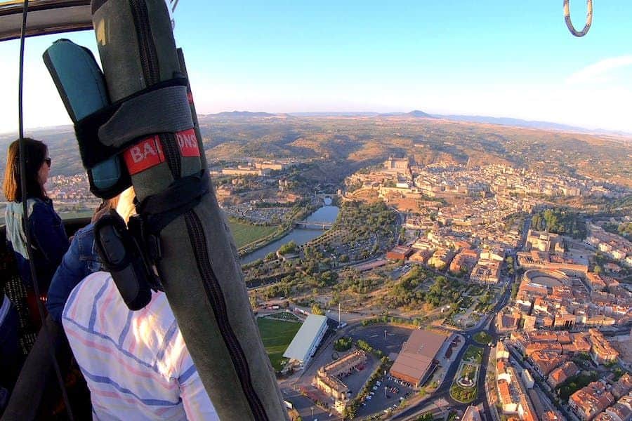 Aerial view of a city with a river, captured from a hot air balloon. The landscape shows buildings, roads, and distant hills.