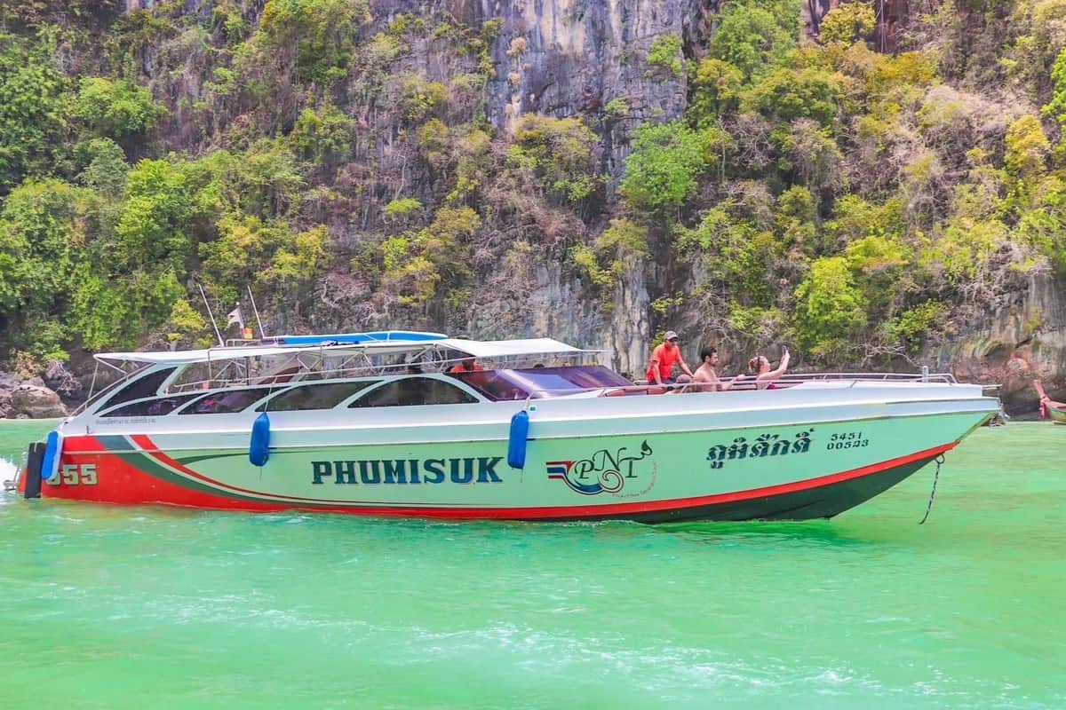 White speedboat named 'PHUMISUK' with people on board cruising past a steep, lush green cliff over emerald water.