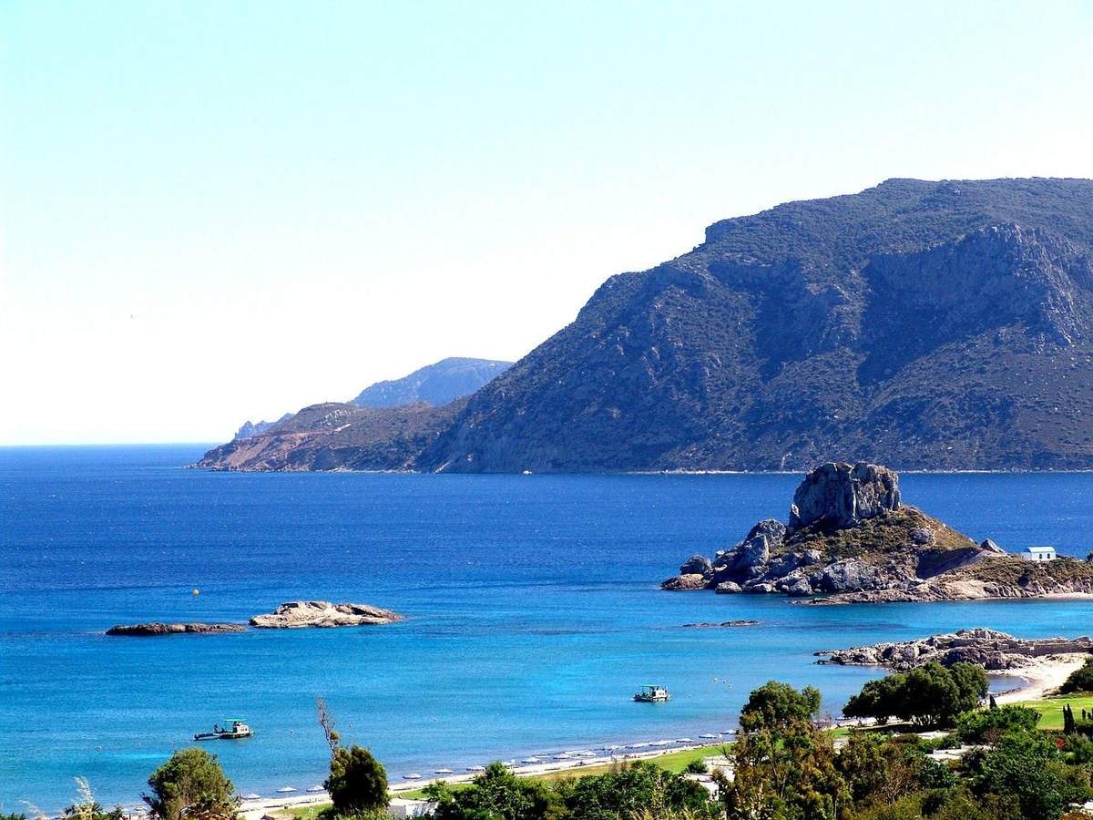 A coastal landscape with blue water, rocky hills in the background, small islands, and trees in the foreground.