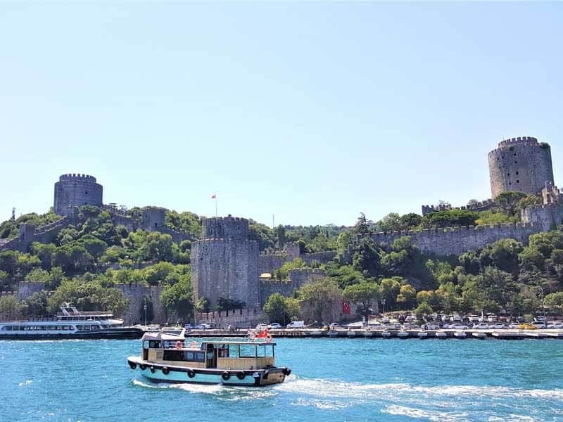 Istanbul: Bosporus & Schwarzes Meer Sightseeing Cruise, Guided Tour + Transport Ein kleines Boot schippert auf einem blauen Fluss vor einer historischen Festung auf einem grünen Hügel unter einem klaren Himmel.