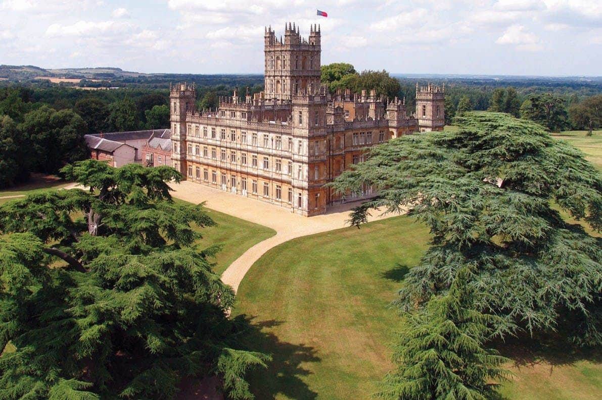A large, ornate castle with towers and a flag on top, surrounded by green lawns and trees under a partly cloudy sky.