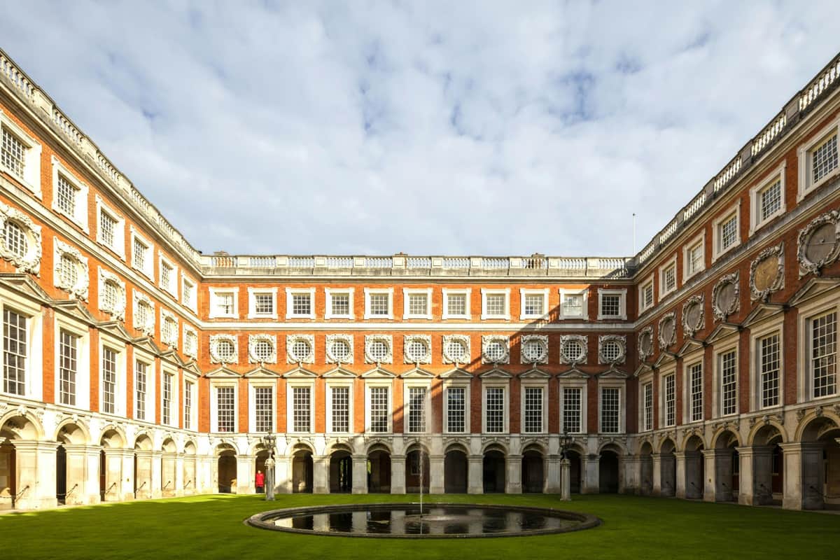 Symmetrical courtyard with a round central fountain, surrounded by a red-brick building with arched windows under a partly cloudy sky.