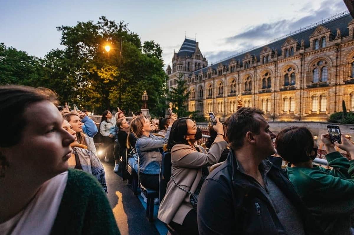 A group of people on an open-top bus tour observing and taking photos near a historic building at dusk.