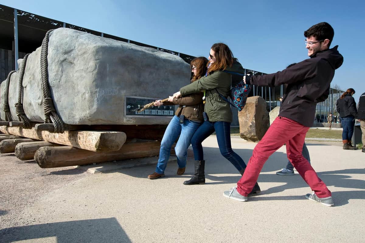Tres personas tiran de una cuerda al aire libre, sonriendo. Al fondo, una gran roca y una valla. Otras personas a lo lejos.
