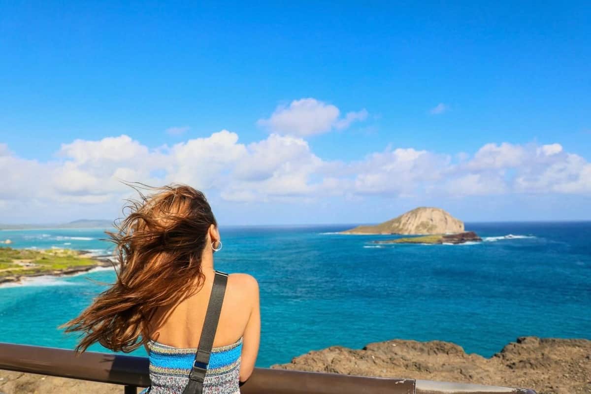 A person with long hair stands on a lookout facing the ocean, with an island in the distance under a partly cloudy sky.