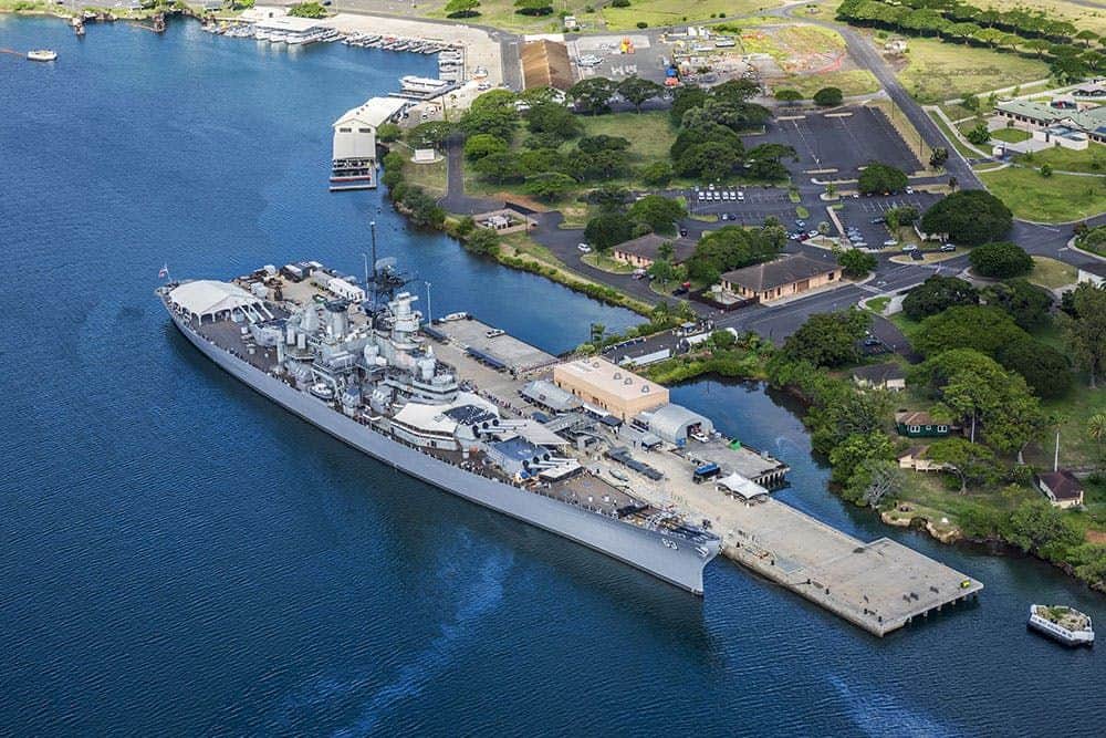 WWII Pearl Harbor Heroes Deluxe Adventure from Honolulu Aerial view of a large battleship docked at a pier surrounded by greenery and buildings near a waterfront.