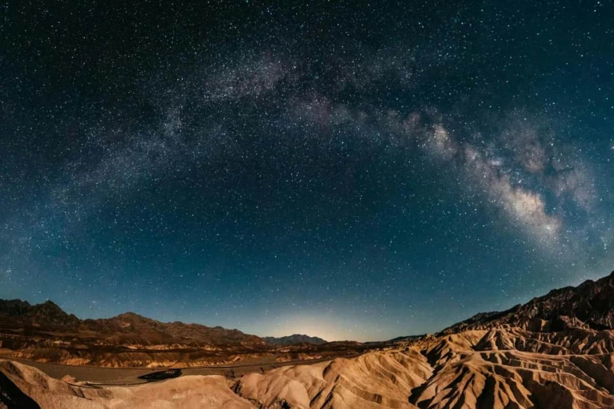 Panoramic night view of a desert landscape with the Milky Way arching across the dark, star-filled sky.