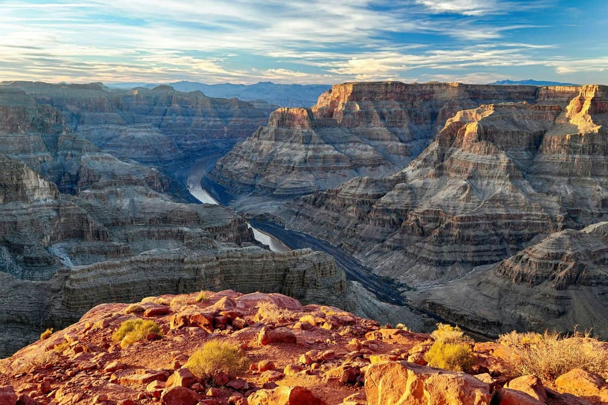 Una vista del Gran Cañón con un río que serpentea por el paisaje rocoso bajo un cielo parcialmente nublado al atardecer.