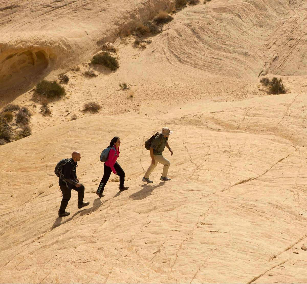 Hiking in the Valley of Fire Three people with backpacks hike up a sandy, rocky slope in a desert landscape with sparse vegetation.