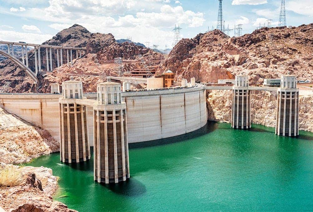A large dam with towers spans a green reservoir, surrounded by rocky terrain, with electrical infrastructure and a blue sky.