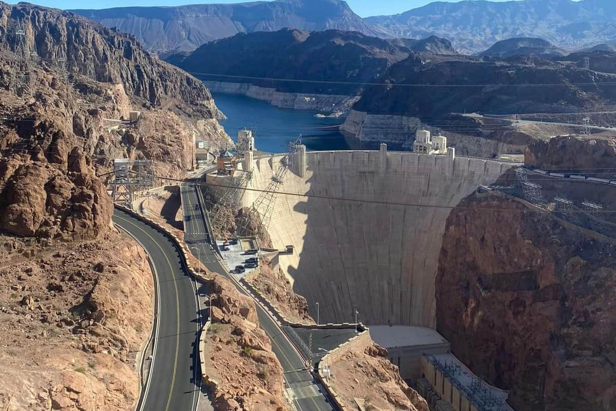 A large dam surrounded by rocky terrain with a road on one side, and a reservoir of water in the background under a clear sky.
