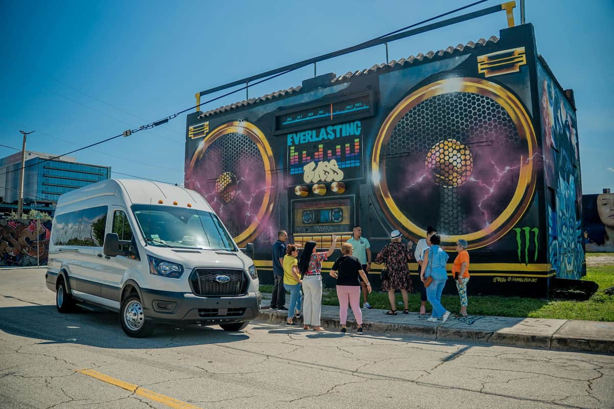 A group of people gathered near a colorful mural of a boombox on a building, with a van parked on the street nearby.