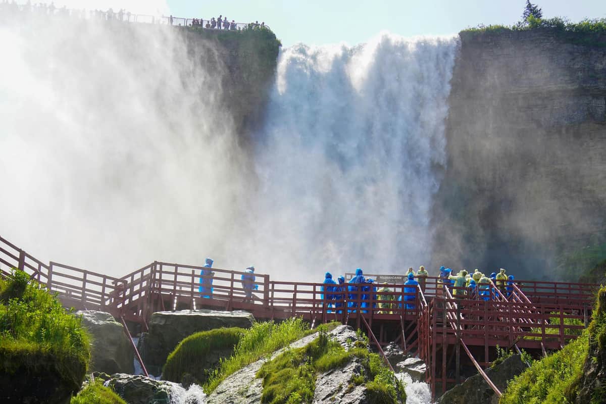 People in blue rain ponchos on a wooden walkway near a large waterfall, with mist and greenery surrounding the area.