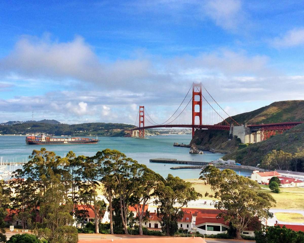 From San Francisco: Day Trip to Muir Woods & Sausalito View of the Golden Gate Bridge with water, trees, and buildings in the foreground under a blue sky with clouds.
