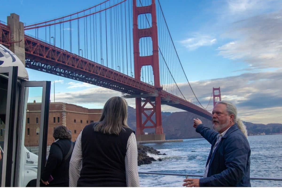 Deux personnes au bord de l'eau, l'une pointant le Golden Gate Bridge, avec un ciel nuageux en arrière-plan.