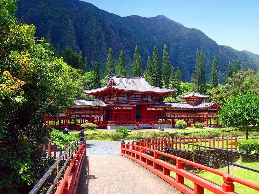 Red Japanese temple with traditional architecture surrounded by lush greenery and mountains, connected by a red arched bridge.