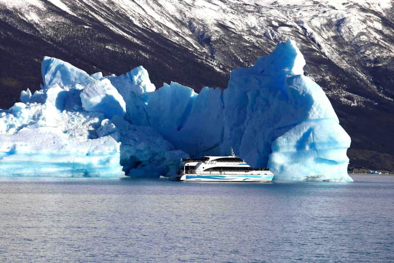 Los Glaciares National Park: Guided Glaciers Boat Tour from El Calafate A boat is near large blue icebergs on a calm body of water, with a forested and snowy mountain in the background.
