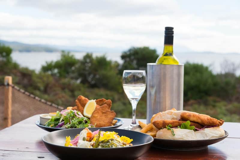 Ein gedeckter Tisch mit Tellern voller Speisen, Brot, Frittiertem, Salat, einer Weinflasche in einer Kühlbox und einem Glas Weißwein.