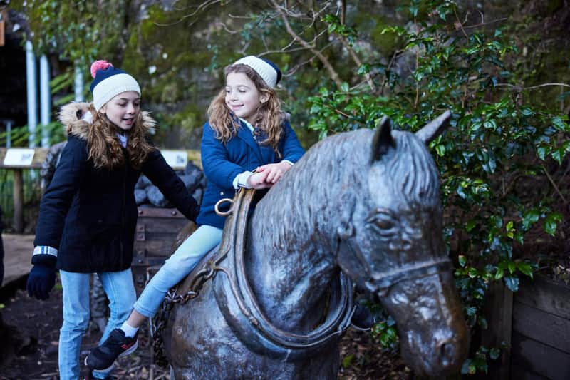 Two children in winter clothing interact with a large sculpture of a horse in an outdoor setting.