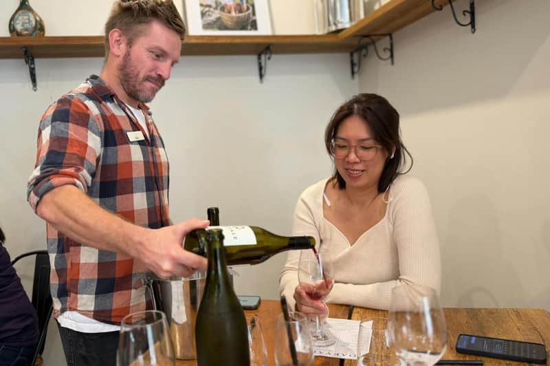 A man pours wine for a woman seated at a table with wine glasses and a tasting sheet in a cozy setting.