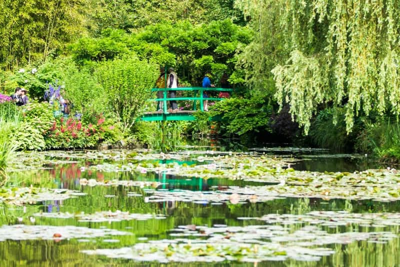 Uno stagno tranquillo con ninfee, circondato dal verde, e un piccolo ponte verde su cui passano delle persone.