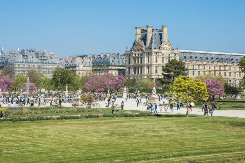 Paris: 1-Day Top Sites Entry + Transfer People walking in a garden with trees, statues, and a historic building in the background under a clear blue sky.