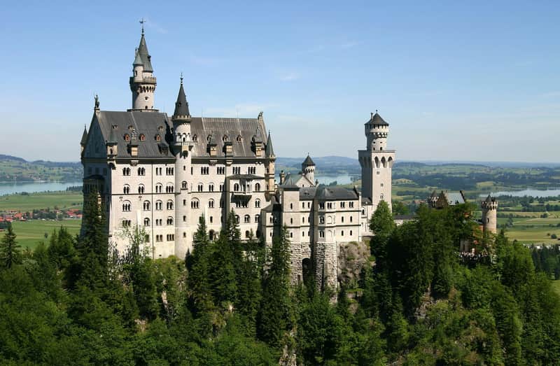 Ein großes, verziertes Schloss mit mehreren Türmen und Türmchen, umgeben von üppigen grünen Bäumen, mit Fernblick auf einen See und die Landschaft.