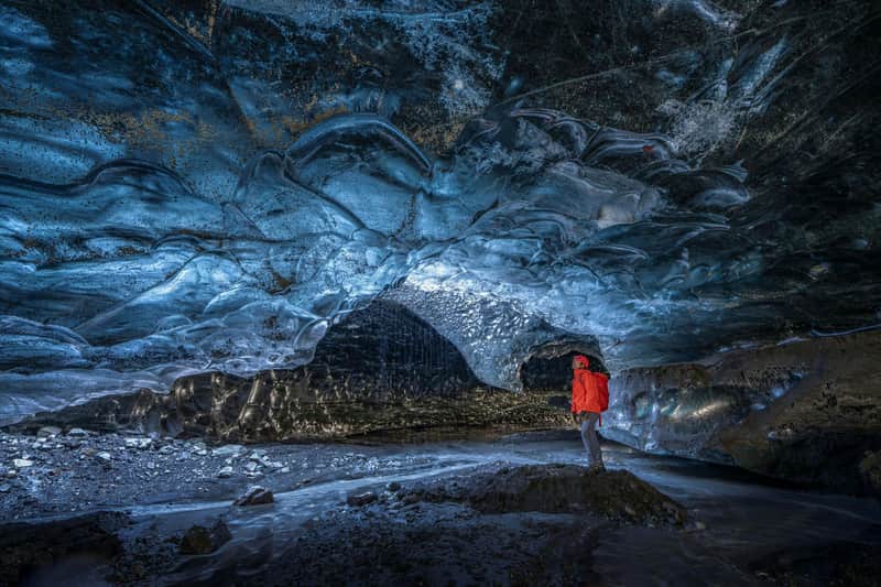 Une personne vêtue d'une veste rouge se tient à l'intérieur d'une grotte de glace d'un bleu éclatant, entourée de parois glacées aux textures variées et d'un sol rocheux.