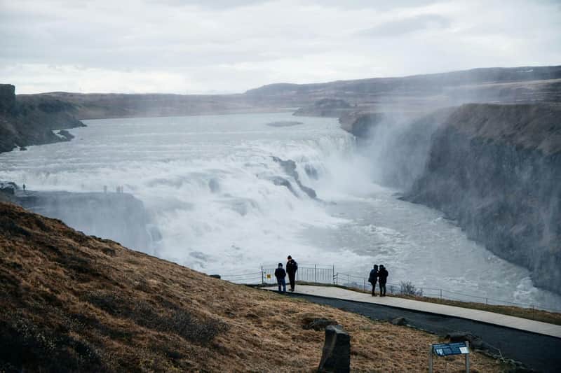 Golden Circle e Langjökull: tour guidato da Reykjavík + giro in motoslitta (Reykjavík) Un gruppo di persone è in piedi vicino a una ringhiera che si affaccia su una grande cascata, in un paesaggio aspro sotto un cielo nuvoloso.