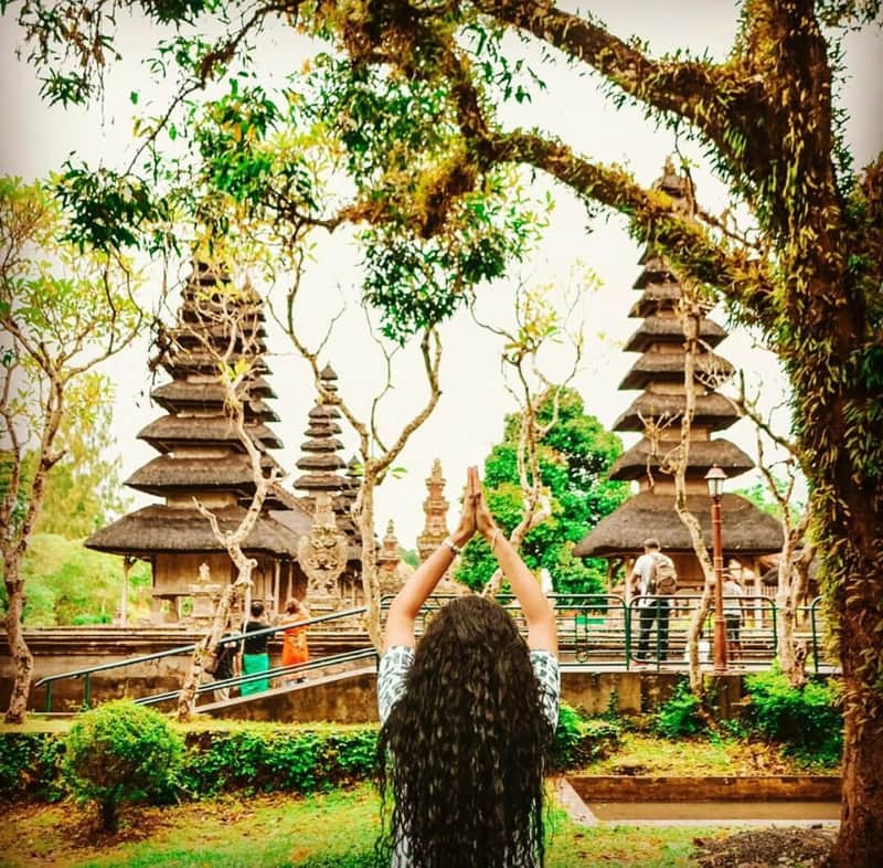 Bali Eat, Pray & Love Private Tour with Guide A person with long, curly hair raising their arms in front of traditional tiered towers in a lush, green temple garden.
