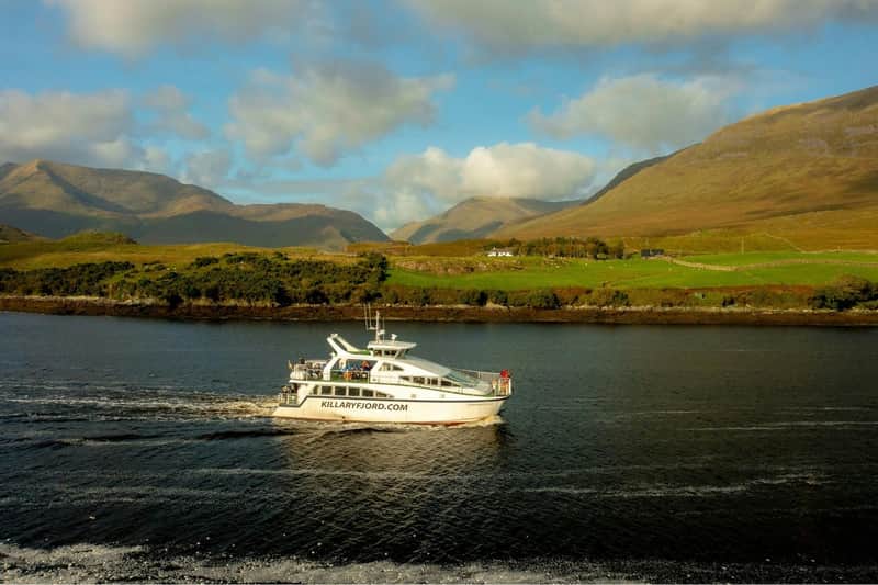 Un barco con la etiqueta "killaryfjord.com" navega por una estrecha masa de agua, con verdes colinas y montañas al fondo.