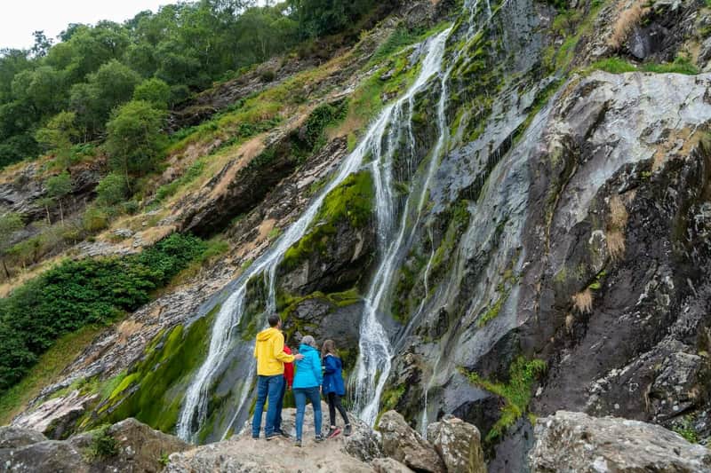Tres personas con chaquetas de colores están de pie sobre unas rocas, admirando una alta cascada cubierta de musgo y rodeada de exuberante vegetación.