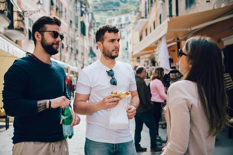 Cinque Terre: Day Tour from Florence Two men in casual clothing stand on a bustling street. One holds a drink and the other holds food. People and buildings in the background.