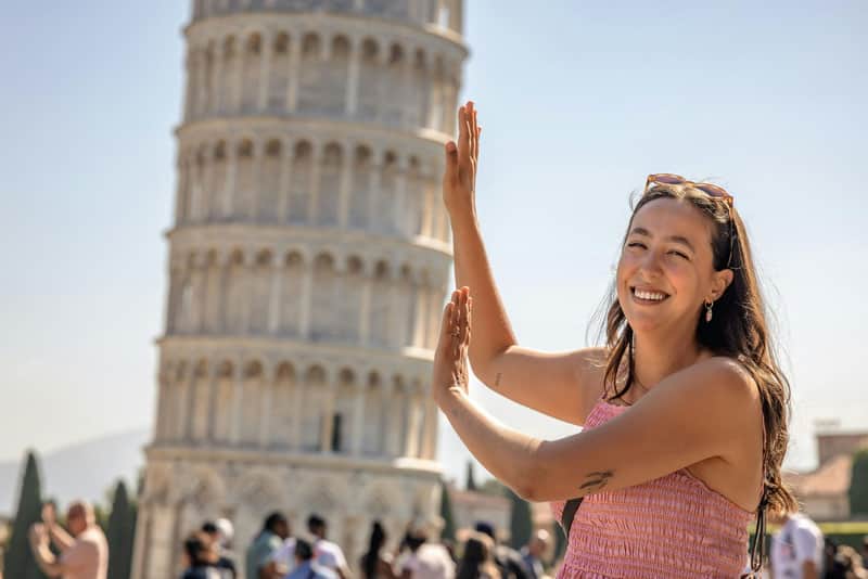 Pisa: Day Trip from Florence A woman smiling and posing with her hands raised in front of the Leaning Tower of Pisa on a sunny day.