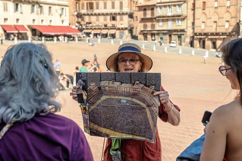 San Gimignano, Siena, Monteriggioni & Chianti: Tour + Lunch + Wine Tasting A person wearing a hat and glasses holds up a poster of a crowded square, surrounded by historic buildings, in an open plaza.