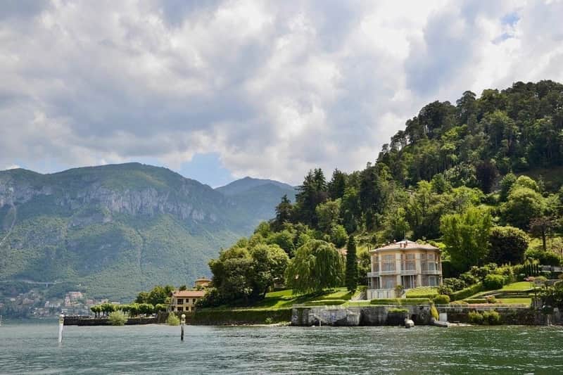 A lakeside villa surrounded by lush greenery with mountains in the background under a partly cloudy sky.