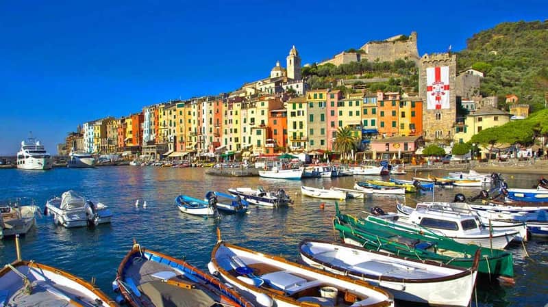 Colorful buildings line a waterfront with numerous boats docked in clear blue water under a bright sky.