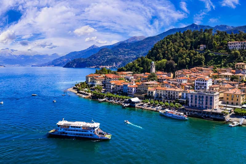 Una vista panoramica di una città sul lago con edifici colorati, barche nell'acqua e montagne sullo sfondo sotto un cielo parzialmente nuvoloso.