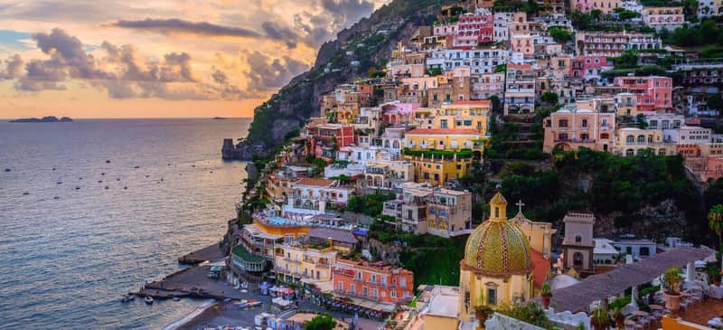 Colorful buildings on a hillside by the sea at sunset, with a gold-domed church in the foreground.