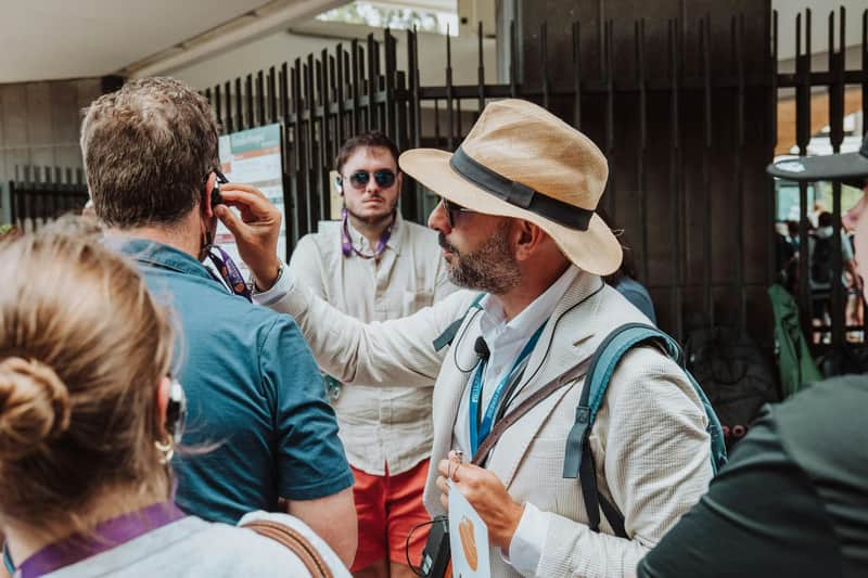 A guide in a light suit and hat adjusts a headset for a tourist in a group, with other people and a fence in the background.