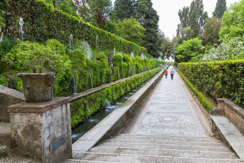 Dos personas caminando por un sendero con fuentes de agua en cascada y exuberante vegetación a la izquierda.