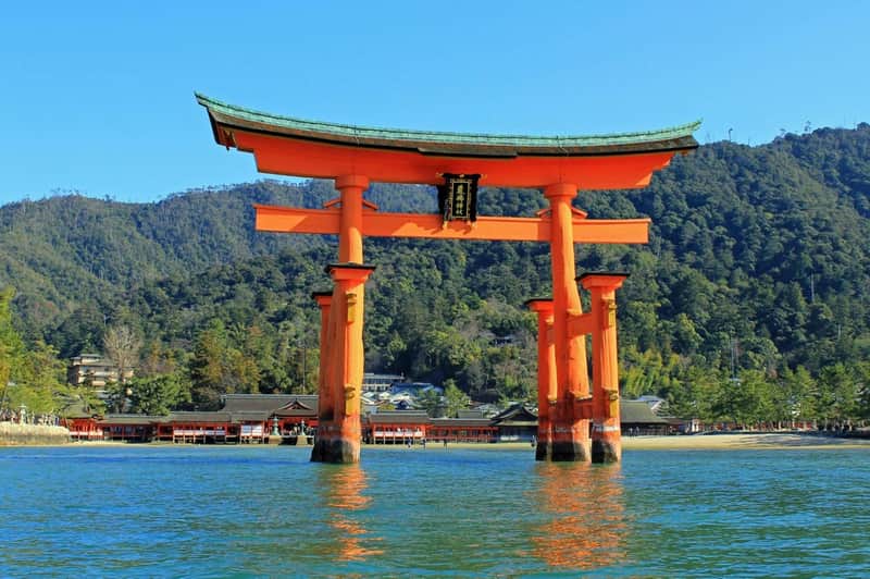 大きな赤い鳥居が水の中に立っており、裏には神社と森の丘が見えます。
