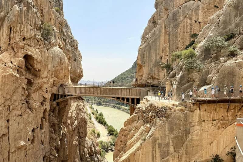 Caminito del Rey: Guided Tour from Málaga + Álora Visit + Lunch A narrow pedestrian bridge spans between two tall rocky cliffs, overlooking a scenic valley and a river below.
