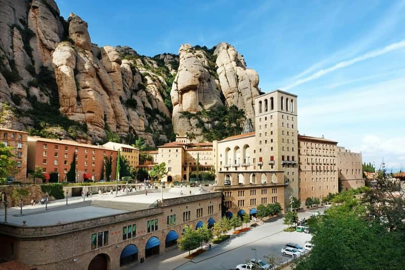 Ensemble de bâtiments historiques avec des arches et une haute tour au pied de montagnes rocheuses et escarpées, sous un ciel bleu et clair.